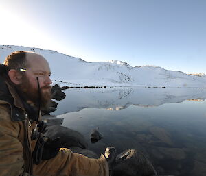 Expeditioner sitting on left hand side of photo looking out at still lake and snow covered hills