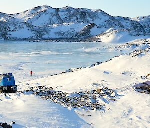 Red field hut and blue tracked vehicle in foreground with frozen fjord and snow covered hills in background