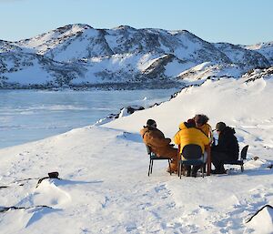 Four expeditioners seated at a table outside in foreground with frozen fjord and hills in background