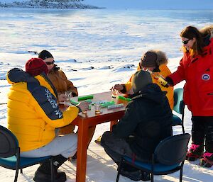 Expeditioner leaning and pouring drinks whilst standing beside a table with four seated expeditioners beside a frozen fjord,