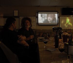 Expeditioner seated at dinner table in foreground with film projected in background