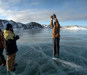 Expeditioner standing over a electric drill with a larger drill piece on a frozen lake