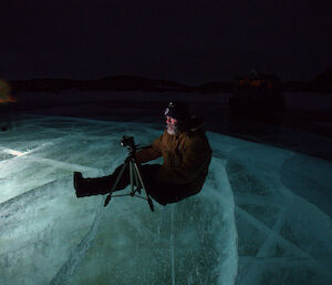 Expeditioner sits atop a frozen lake illuminated by torchlight set into holes in the ice surrounded by darkness
