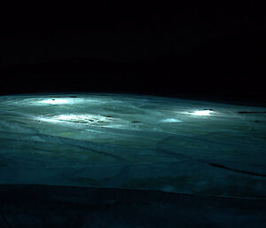 A frozen lake illuminated by torchlight shining a blue green light through the ice.. the darkness of night above