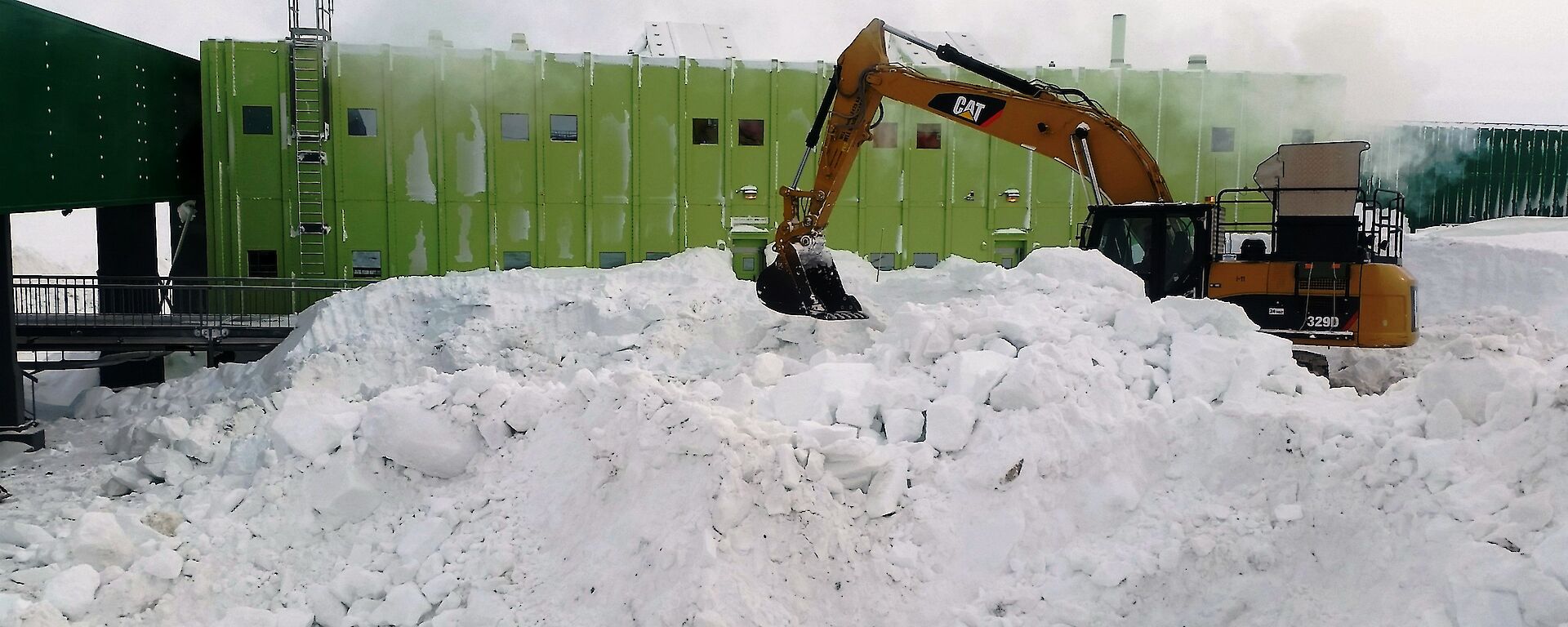 An excavator clears snow in foreground with a large green building in background