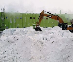 An excavator clears snow in foreground with a large green building in background