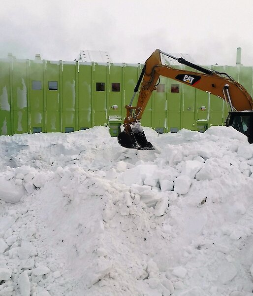 An excavator clears snow in foreground with a large green building in background