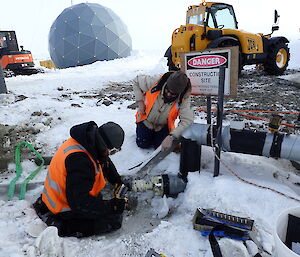 Two expeditioners kneeling in front of exposed site services piping