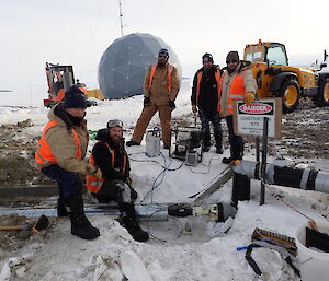 Five expeditioners standing above exposed pipe with the ANARESAT comms dome in background