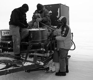 Three expeditioners standing beside and sitted atop a trailer carrying fuel drums.. holding a fuel pump