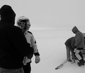 Expeditioner facing camera in foreground behind an expeditioner not facing camera. In background other expeditioners beside a trailer parked on a white icy surface