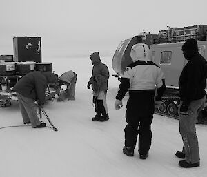 Expeditioners standing in foreground right, two securing a sled with fuel drums on it. The trailer of the second tracked vehicle in background