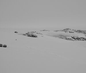 Landscape photo of a tracked vehicle in distance parked on a long slope with expeditioners inspecting an access track down onto a frozen lake. snow covered hills and rocky outcrops with sheer ice walls in background