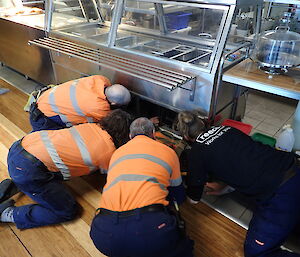 Four expeditioners kneeling with heads converging beneath a refrigerated cabinet
