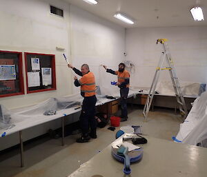 Two expeditioners standing with paint rollers facing camera in profile as they paint walls white with a third expeditioner lying under the bench