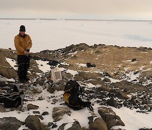 Expeditioner standing beside a remote camera on rocky terrain with open water and a very large ice ice berg