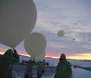 Expeditioners in flame retardant clothing in a line to release large white weather balloons off a snow covered ramp