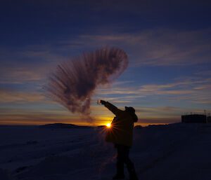 Expeditioner silhouetted against sun on horizon whilst throwing water that freezes in mid air