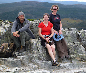 Three adults facing camera smiling sitting on rocks with dark mountains in background