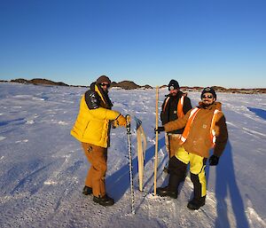 Three expeditioners facing camera standing on snow with a broken bamboo cane protruding from the snow.