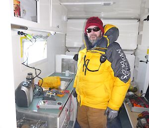 Expeditioner facing camera inside a small accommodation unit with electrical equipment beside him