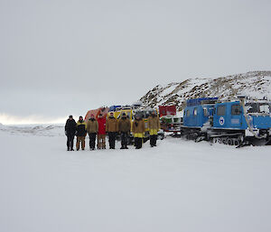 8 expeditioners stand in a line in mock formal fashion in front of some tracked vehicles