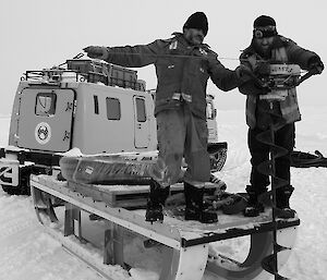 Two expeditioners stand on a sledge preparing to drill a hole using an auger