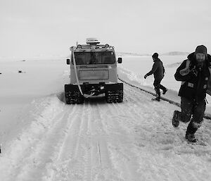 Expeditioners standing beside water hoses on the right hand side of a snow road with a tracked vehicle in background