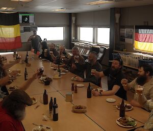 A large group of expeditioners around a table with german cuisine on the table and german flags hanging from the ceiling