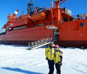 Two expeditioners standing on sea ice next to the hull of an orange icebreaker