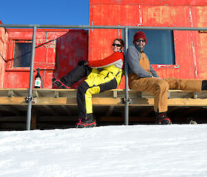 Two expeditioners seated back to back on the deck of a field hut