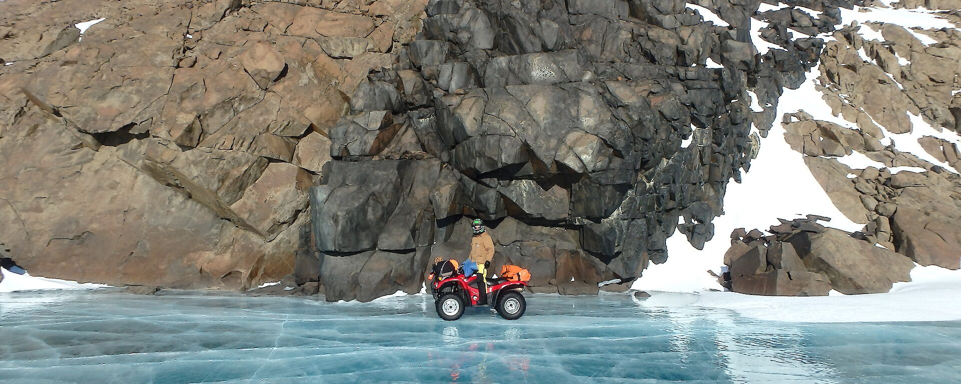 Expeditioner standing on a quad bike above a frozen lake with rocky cliff in background