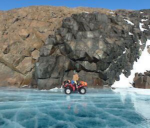 Expeditioner standing on a quad bike above a frozen lake with rocky cliff in background