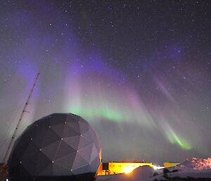 A green and blue aurora hanging over the ANARESAT dome next to the operations building at night