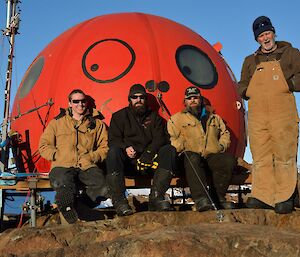 Four expeditioners perched in front of a red field hut facing the camera
