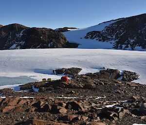 Red field hut and yellow tracked vehicle parked below it with ice and rock filled landscape in background