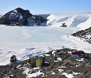 A series of field huts located on a rocky outcrop with frozen sea ice and a large ice cliff in background