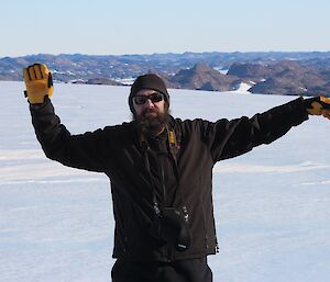 Expeditioner standing on an icy plateau with arms outstretched. Brown hills in background