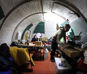 Two expeditioners inside a field hut with equipment and pots strewn around