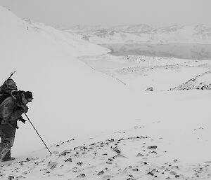 Expeditioner in foreground resting on a walking stick with snow covered landscape and lake in background