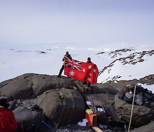 Expeditioners standing on a rocky outcrop holding a red ensign