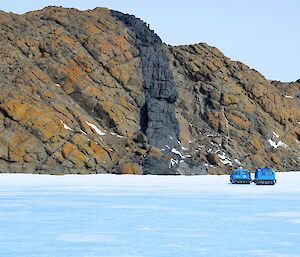 Blue tracked vehicle driving upon sea ice beside large rocky outcrop