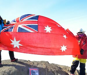 Two expeditioners standing beside a large flag with rocky terrain in background