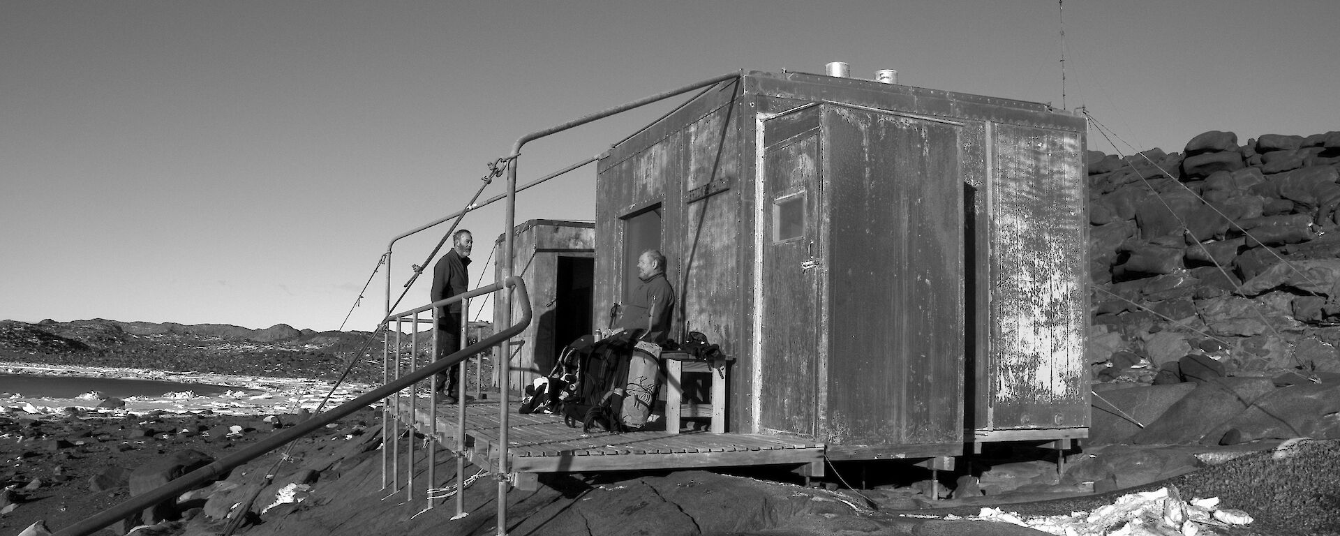 Two expeditioners outside on the sun deck of a hut with rocky terrain in the background