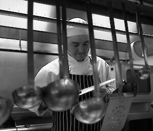 Expeditioner standing behind a row of hanging ladles and kitchen utensils sharpening a knife