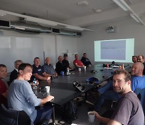 A group of people sit in a classroom with a digital display screen