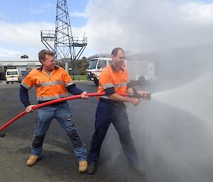 Two men in trades overalls hold a high pressure fire fighting hose