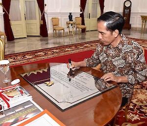 The Indonesian President siting at a desk signing a plaque