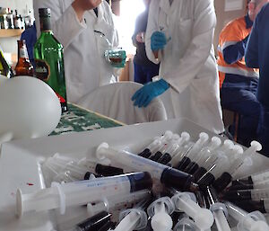 Two scientists in white coats standing behind a tray of plastic syringes used as props