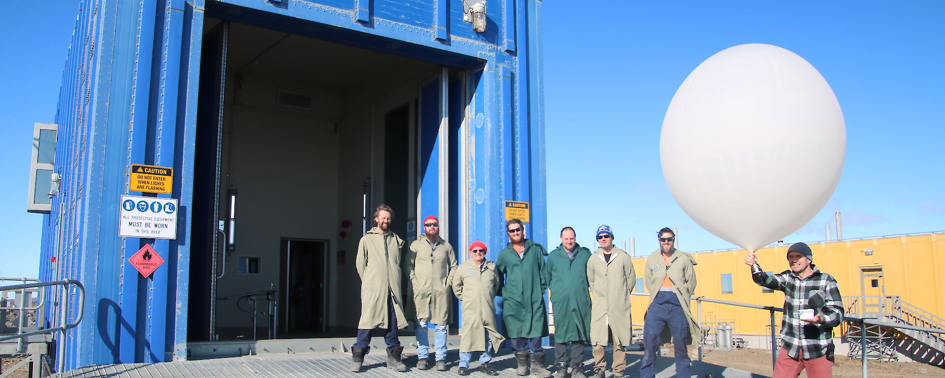 Expeditioners stand outside of a shipping container office, one holds a large balloon